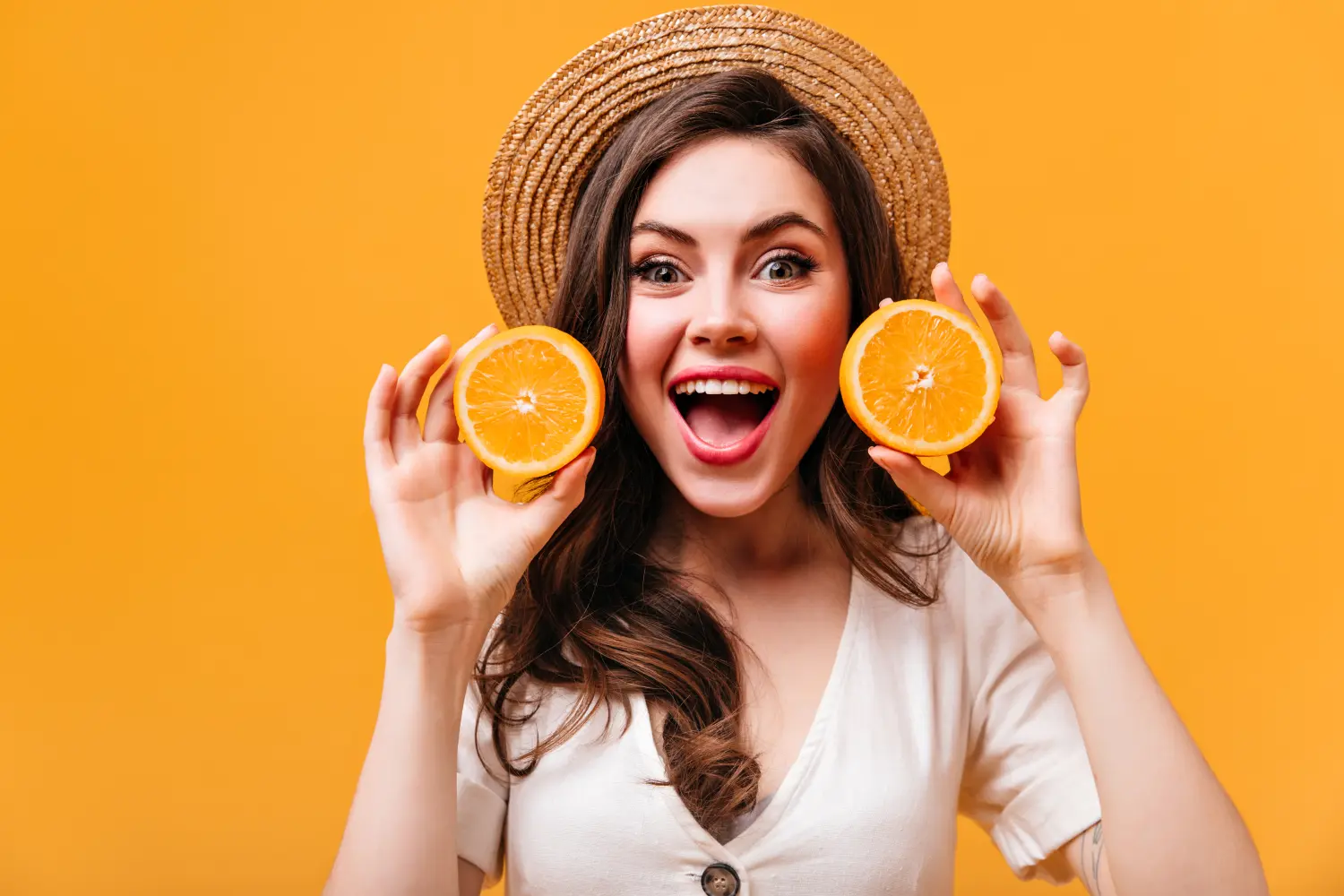 Charming woman with green eyes looks into camera with delight and holds oranges on isolated background.